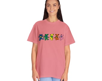 Puede incluir: Una camiseta rosa con una fila de cinco osos bailando en colores arcoíris. Los osos están delineados en negro y tienen un diseño simple.