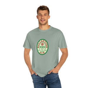 May include: A light green t-shirt featuring a vintage-style logo for Ballantine Ale. The logo is a green circle with a red star on each side and the words "Ballantine Ale" in red lettering. Inside the circle is a red "XXX" with a green clover design.