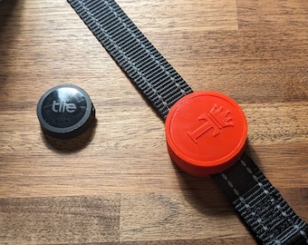 May include: A black and gray dog collar with a silver buckle and a red circular attachment with a white emblem. A small black Tile tracker is next to the collar on a wooden surface.