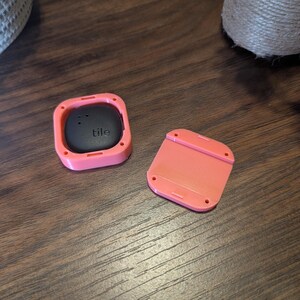 May include: A black "tile" tracker in a coral-colored square case, with a matching detached cover. The tracker has a smooth, rounded surface with the brand name printed on it. The case has a square shape with rounded corners.