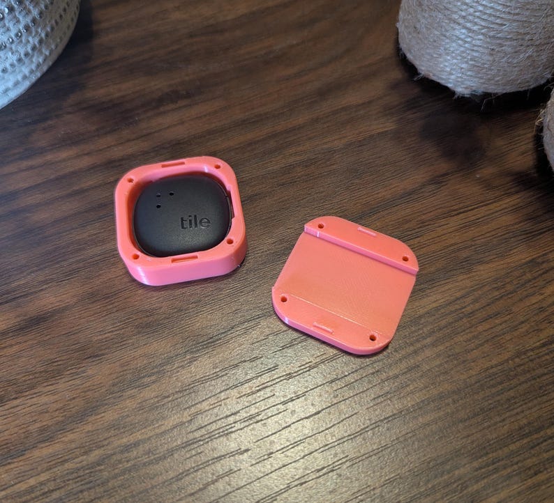 May include: A black "tile" tracker in a coral-colored square case, with a matching detached cover. The tracker has a smooth, rounded surface with the brand name printed on it. The case has a square shape with rounded corners.