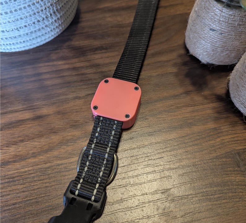 May include: Close-up of a black and gray dog collar with a peach-colored square component. The collar has a metal D-ring and a black plastic buckle. The square component is secured with four black screws.