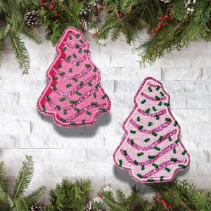 May include: Two pink Christmas tree-shaped patches with glitter and green sprinkles. The patches have a stitched design and are set against a white brick wall with evergreen branches and pinecones.