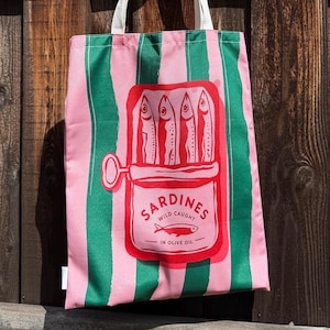 May include: A tote bag featuring vertical pink and green stripes with a central red graphic of an open sardine tin. The graphic depicts four fish inside the tin with the text SARDINES WILD CAUGHT IN OLIVE OIL and a small fish illustration.