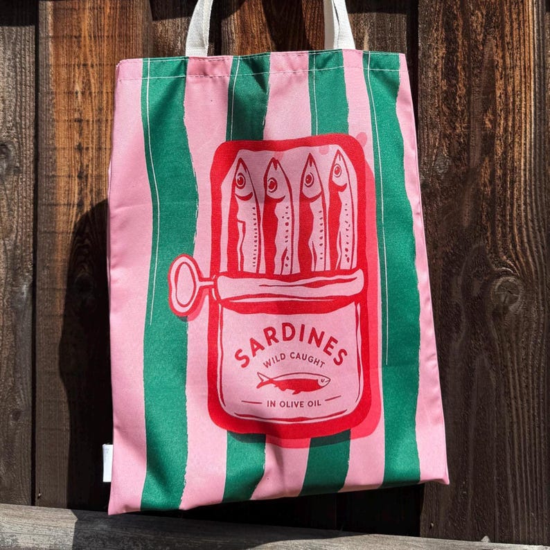 May include: A tote bag featuring vertical pink and green stripes with a central red graphic of an open sardine tin. The graphic depicts four fish inside the tin with the text SARDINES WILD CAUGHT IN OLIVE OIL and a small fish illustration.