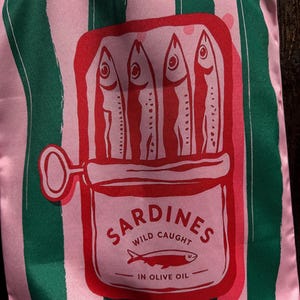 May include: A fabric bag featuring vertical pink and dark green stripes. The center displays a red graphic of an open sardine tin containing four fish. The text reads SARDINES WILD CAUGHT IN OLIVE OIL with a small fish icon below the text.