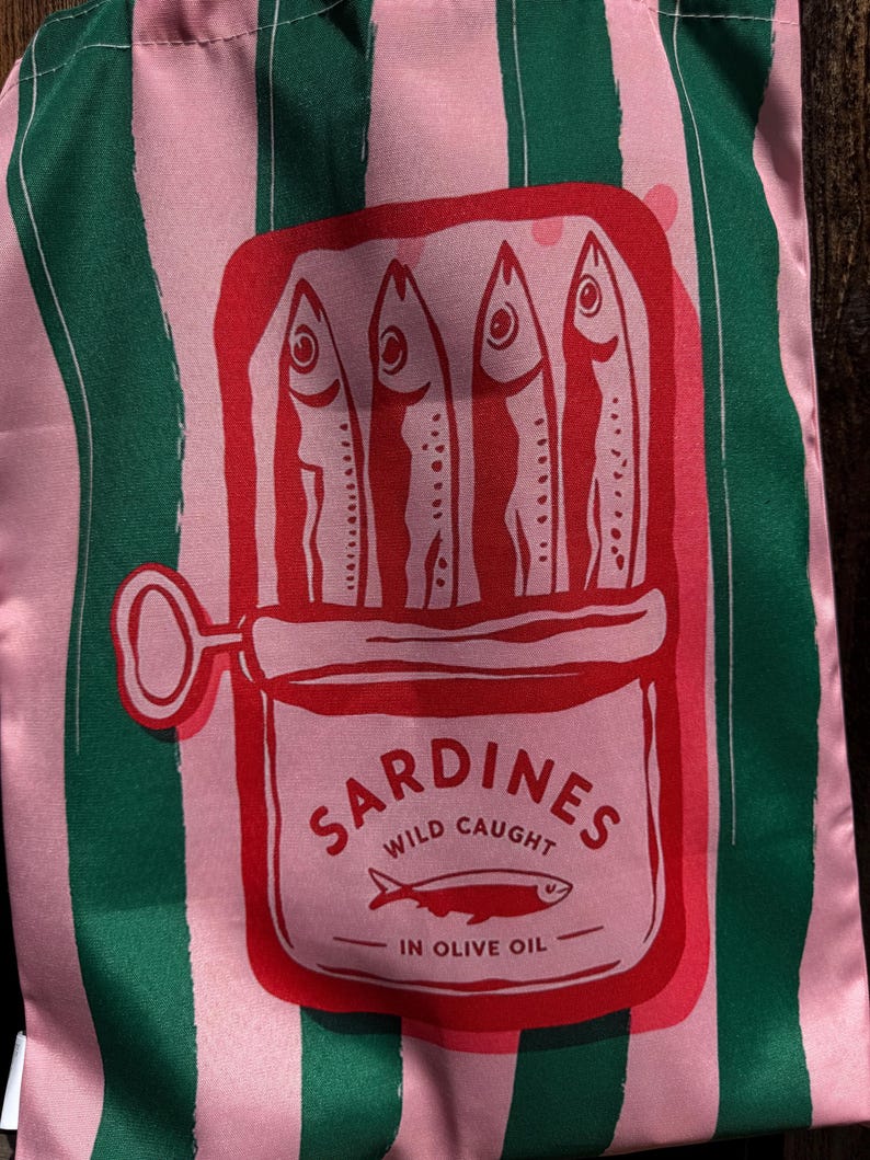 May include: A fabric bag featuring vertical pink and dark green stripes. The center displays a red graphic of an open sardine tin containing four fish. The text reads SARDINES WILD CAUGHT IN OLIVE OIL with a small fish icon below the text.