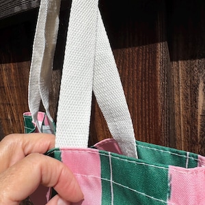 May include: A tote bag featuring a vertical striped pattern in alternating shades of forest green and light pink, accented by thin white pinstripes. The bag includes sturdy, off-white woven fabric handles attached to the top edge with visible stitching.
