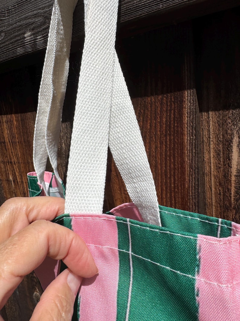 May include: A tote bag featuring a vertical striped pattern in alternating shades of forest green and light pink, accented by thin white pinstripes. The bag includes sturdy, off-white woven fabric handles attached to the top edge with visible stitching.