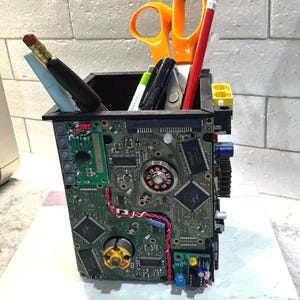 May include: A square pencil holder constructed from recycled computer circuit boards and electronic components. The exterior features green printed circuit boards, integrated chips, and copper coils. It contains orange scissors, a red pencil, and pens.