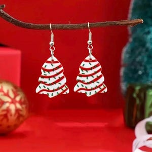 May include: A pair of Christmas tree earrings. Each earring is shaped like a white Christmas tree with red stripes and green sprinkles. The earrings hang from silver-colored fish hook ear wires.