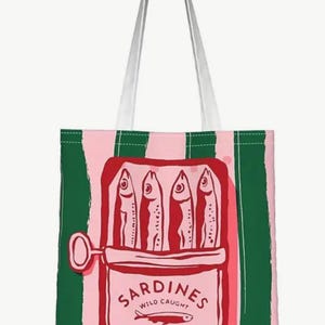 May include: A tote bag with vertical pink and dark green stripes featuring a central graphic of an open red tin of sardines. The illustration shows four fish inside the tin with the text SARDINES WILD CAUGHT IN OLIVE OIL and a small fish icon.