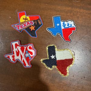 May include: Four embroidered patches shaped like the state of Texas. One features a cowboy silhouette, sunset, and the word TEXAS. Another shows the Texas flag. A third displays the word TEXAS in white letters. The fourth is a textured Texas flag.