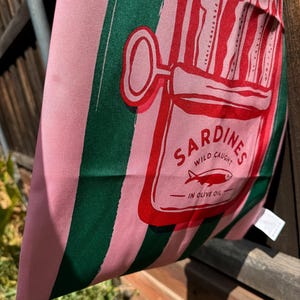 May include: A pink and dark green striped fabric tote bag featuring a red graphic of an open sardine tin. The print includes the text SARDINES, WILD CAUGHT, and IN OLIVE OIL, with an illustration of three fish inside the rectangular tin container.