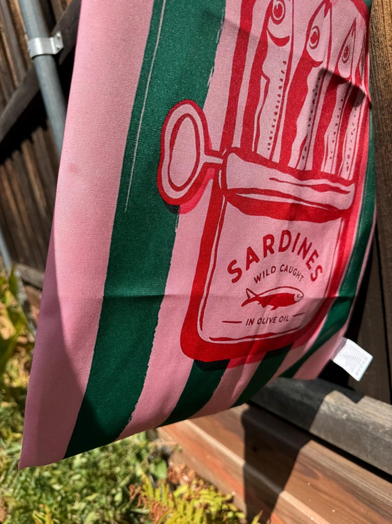 May include: A pink and dark green striped fabric tote bag featuring a red graphic of an open sardine tin. The print includes the text SARDINES, WILD CAUGHT, and IN OLIVE OIL, with an illustration of three fish inside the rectangular tin container.