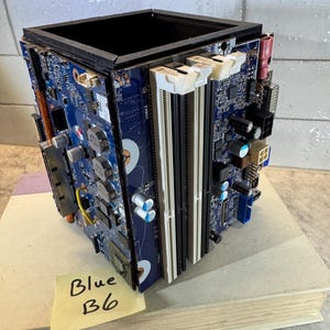 May include: A square-shaped desk organizer constructed from recycled blue computer motherboards. The structure features exposed electronic components, including white RAM slots, capacitors, and various ports, with a black frame at the top edge.
