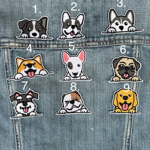May include: A collection of nine embroidered dog patches, each featuring a different breed. The patches are in various colors, including black, white, brown, and yellow, and are affixed to a denim jacket. Each dog is peeking over a horizontal line.