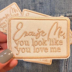 May include: Cream-colored embroidered patch with the text "Excuse Me, you look like you love me" in a cursive and block font. The patch has a gold border and is rectangular. Several patches are visible.