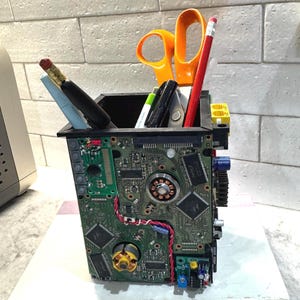 Peut inclure: Un pot à crayons carré fabriqué à partir de circuits imprimés verts recyclés et de composants électroniques. L'extérieur présente des puces, un moteur et des fils visibles. Il contient des ciseaux à poignées orange, un crayon rouge et des stylos.