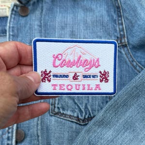 May include: White embroidered patch with blue trim featuring the text "Cowboys" in pink lettering, "The Legend & Since 1973" in smaller pink lettering, and "TEQUILA" in pink lettering below. The patch features two red and white lion emblems on either side of the text.