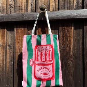 May include: A pink and green striped tote bag featuring a central graphic of an open tin of sardines. The red illustration depicts four fish inside the tin with the text SARDINES WILD CAUGHT IN OLIVE OIL and a small fish icon below.