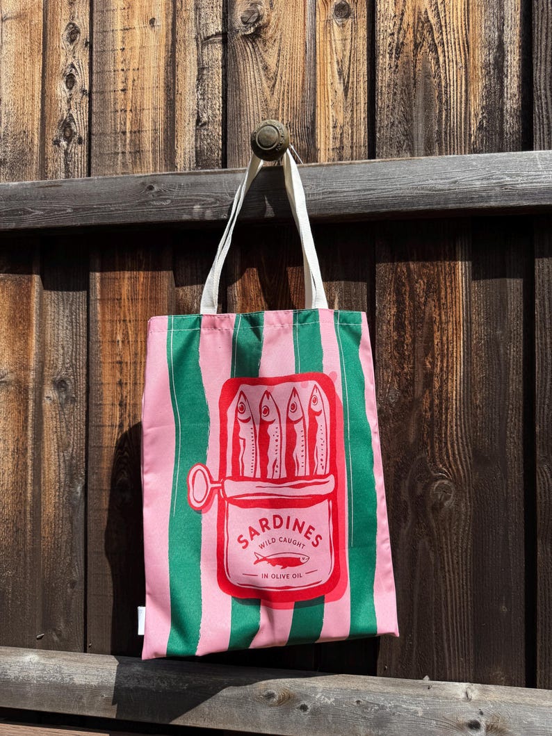 May include: A pink and green striped tote bag featuring a central graphic of an open tin of sardines. The red illustration depicts four fish inside the tin with the text SARDINES WILD CAUGHT IN OLIVE OIL and a small fish icon below.