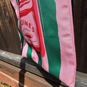 May include: A fabric tote bag featuring vertical pink and dark green stripes. The center graphic depicts a red rectangular sardine tin label with the text SARDINES, WILD CAUGHT, and IN OLIVE OIL, accompanied by a small fish illustration.
