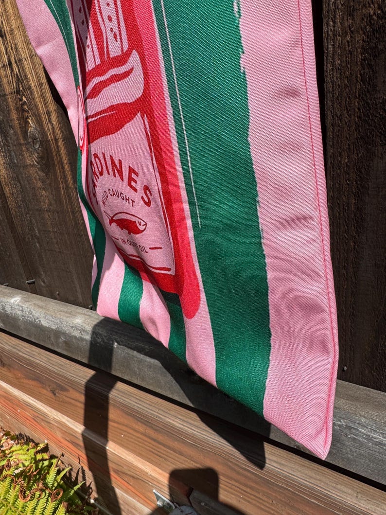May include: A fabric tote bag featuring vertical pink and dark green stripes. The center graphic depicts a red rectangular sardine tin label with the text SARDINES, WILD CAUGHT, and IN OLIVE OIL, accompanied by a small fish illustration.
