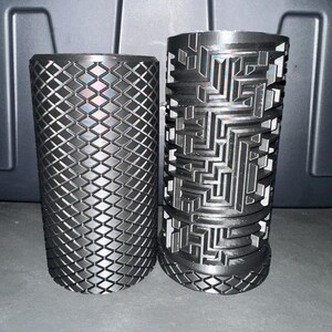 May include: Two black cylindrical objects with intricate geometric patterns. One features a diamond grid design, while the other showcases a maze-like structure. Both are likely decorative or functional items.