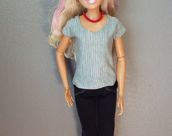 May include: A doll with blonde hair streaked with pink, wearing a light blue striped V-neck shirt, black pants, and a red beaded necklace. The doll has blue eyes and is smiling.