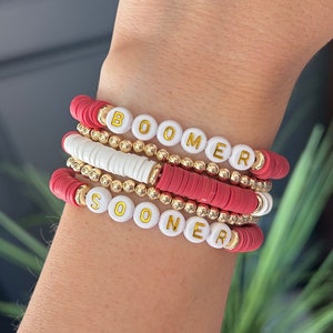 Puede incluir: Un conjunto de pulseras de cuentas rojas, blancas y doradas. Las pulseras muestran las palabras "BOOMER" y "SOONER" en letras doradas sobre cuentas blancas. Las pulseras combinan cuentas doradas redondas con cuentas rojas y blancas planas.