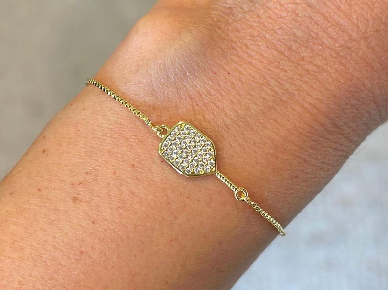 May include: A gold-colored bracelet with a paddle-shaped charm. The charm is encrusted with small, clear crystals. The bracelet is on a person's wrist.