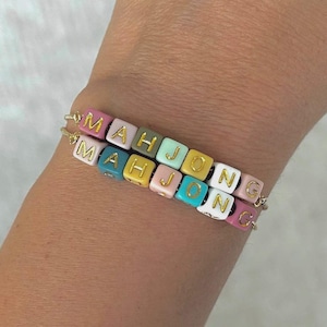 May include: Two stacked bracelets featuring square beads in various pastel colors including pink, olive green, light blue, yellow, and white. Each bead displays a gold letter, with the beads collectively spelling out the word MAHJONG on both strands.