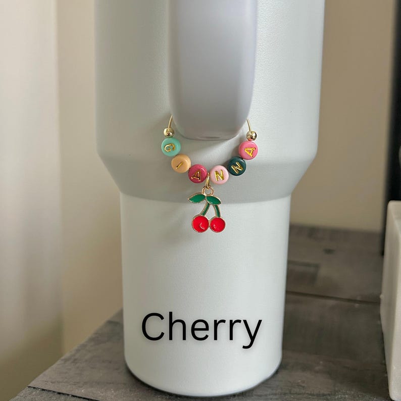 May include: A gold-colored charm with colorful beads spelling "JANN" and a red cherry pendant. The charm is attached to a white tumbler. The word "Cherry" is printed in black at the bottom of the tumbler.