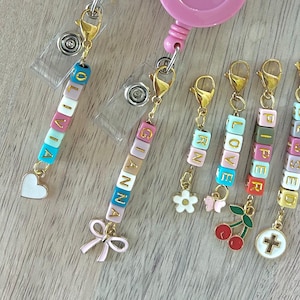 May include: A collection of personalized badge reels with gold-colored hardware. Each reel features a name spelled out with colorful letter beads. Charms include a heart, bow, flower, butterfly, cherries, and a cross.