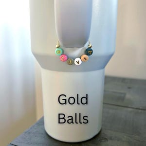 May include: A light gray tumbler with a gold hoop charm. The charm features colorful beads spelling "LOVE". The tumbler has the words "Gold Balls" printed on it in black.