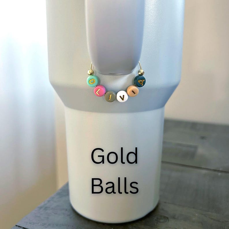May include: A light gray tumbler with a gold hoop charm. The charm features colorful beads spelling "LOVE". The tumbler has the words "Gold Balls" printed on it in black.