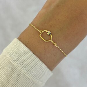 May include: A gold-colored bracelet featuring a geometric charm with a small black and white beaded accent. The bracelet is worn on a wrist, and the chain has a delicate, textured design.