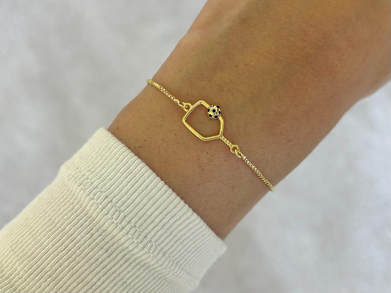 May include: A gold-colored bracelet featuring a geometric charm with a small black and white beaded accent. The bracelet is worn on a wrist, and the chain has a delicate, textured design.