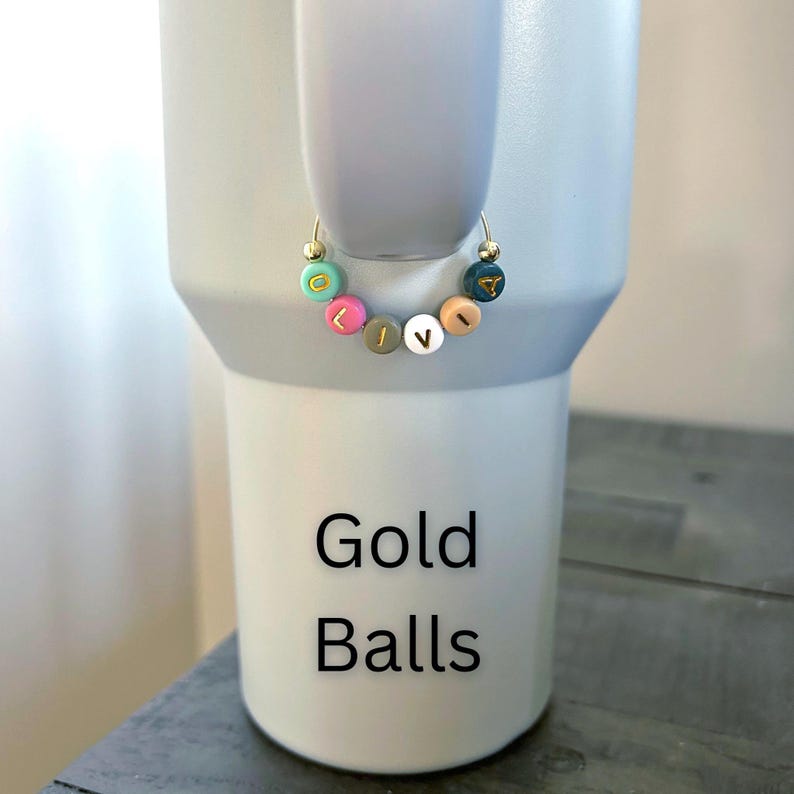 May include: A gold hoop earring with colorful letter beads spelling "LOVE" is displayed. The earring is shown against a light gray tumbler. The tumbler has the words "Gold Balls" printed on it.