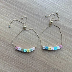 May include: Two gold-tone chain bracelets with adjustable sliding clasps. Each bracelet features a row of square, pastel-colored beads spelling out the word MAHJONG in gold letters. The beads are pink, yellow, blue, and white, with a small crystal accent.
