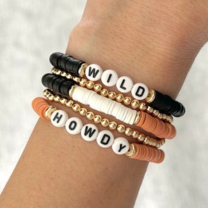 May include: A set of stacked bracelets with gold beads and letter beads spelling out "HOWDY" and "WILD". The bracelets are in shades of orange, black, and white.
