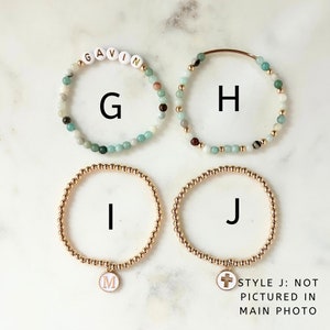 May include: Four bracelets with different charms. The first bracelet is made of light blue and green beads with the name "GAVIN" spelled out in white letters. The second bracelet is made of light blue and green beads with a gold clasp. The third bracelet is made of gold beads with a white charm that has the letter "M" on it. The fourth bracelet is made of gold beads with a white charm that has a cross on it. The text "STYLE J: NOT PICTURED IN MAIN PHOTO" is written below the bracelets.