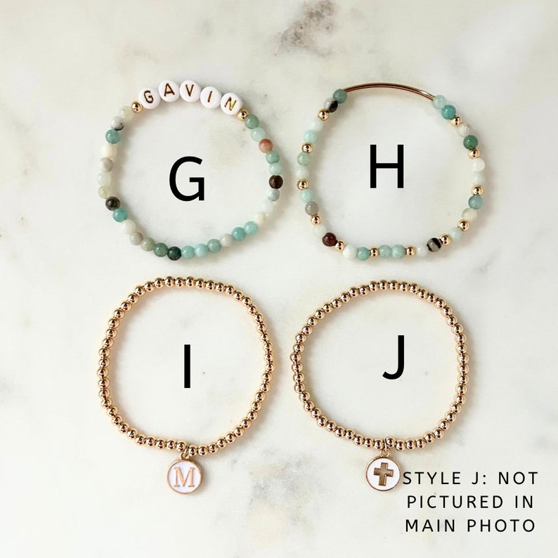 May include: Four bracelets with different charms. The first bracelet is made of light blue and green beads with the name "GAVIN" spelled out in white letters. The second bracelet is made of light blue and green beads with a gold clasp. The third bracelet is made of gold beads with a white charm that has the letter "M" on it. The fourth bracelet is made of gold beads with a white charm that has a cross on it. The text "STYLE J: NOT PICTURED IN MAIN PHOTO" is written below the bracelets.