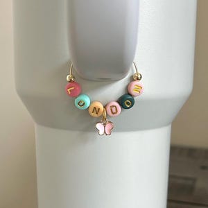 May include: A gold-colored hoop charm with colorful letter beads spelling "LONDON" and a small pink butterfly charm. The beads are in various colors, including pink, teal, orange, and green, with gold lettering. The charm is attached to a white tumbler.