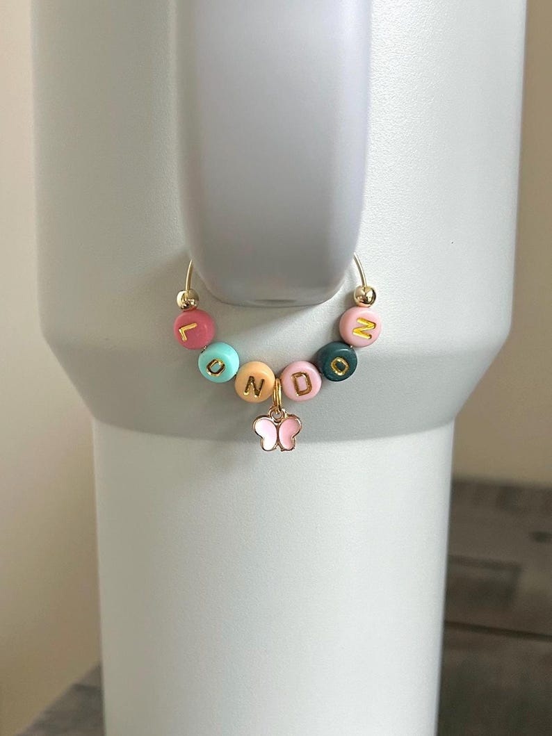 May include: A gold-colored hoop charm with colorful letter beads spelling "LONDON" and a small pink butterfly charm. The beads are in various colors, including pink, teal, orange, and green, with gold lettering. The charm is attached to a white tumbler.