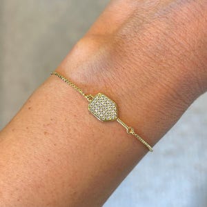 May include: A gold-colored bracelet with a shield-shaped pendant. The pendant is encrusted with small, clear crystals. The bracelet is on a delicate chain and is worn on a person's arm.