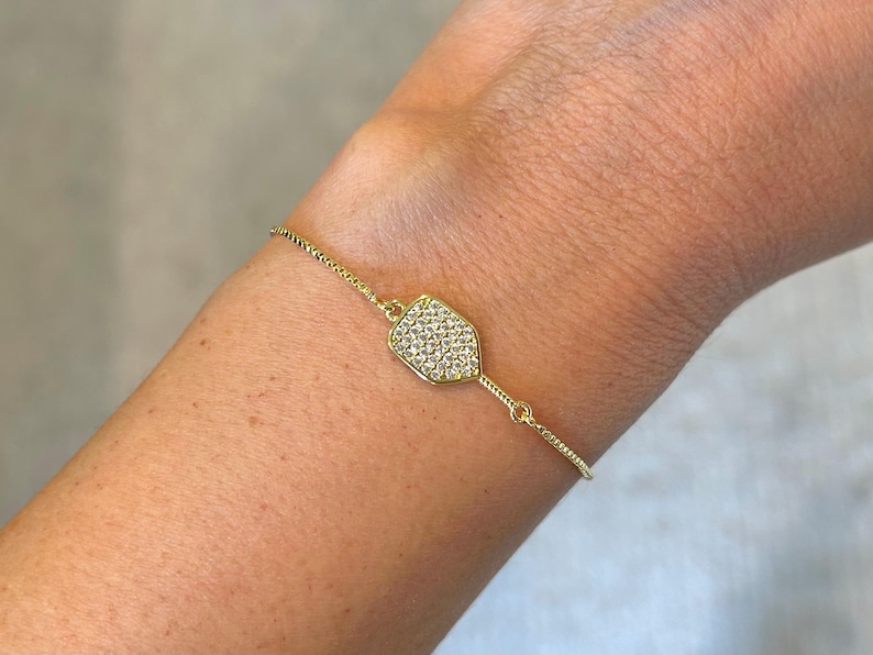 May include: A gold-colored bracelet with a shield-shaped pendant. The pendant is encrusted with small, clear crystals. The bracelet is on a delicate chain and is worn on a person's arm.