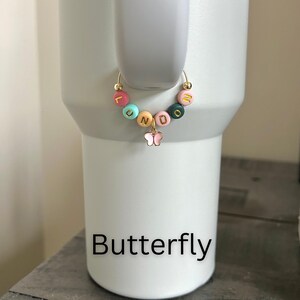 May include: A gold-colored hoop earring with colorful letter beads spelling "LUNDO" and a small pink butterfly charm. The earring is displayed on a white tumbler. The word "Butterfly" is printed in black at the bottom of the image.