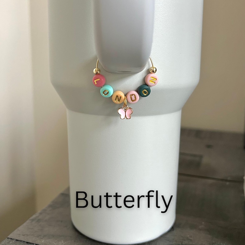 May include: A gold-colored hoop earring with colorful letter beads spelling "LUNDO" and a small pink butterfly charm. The earring is displayed on a white tumbler. The word "Butterfly" is printed in black at the bottom of the image.